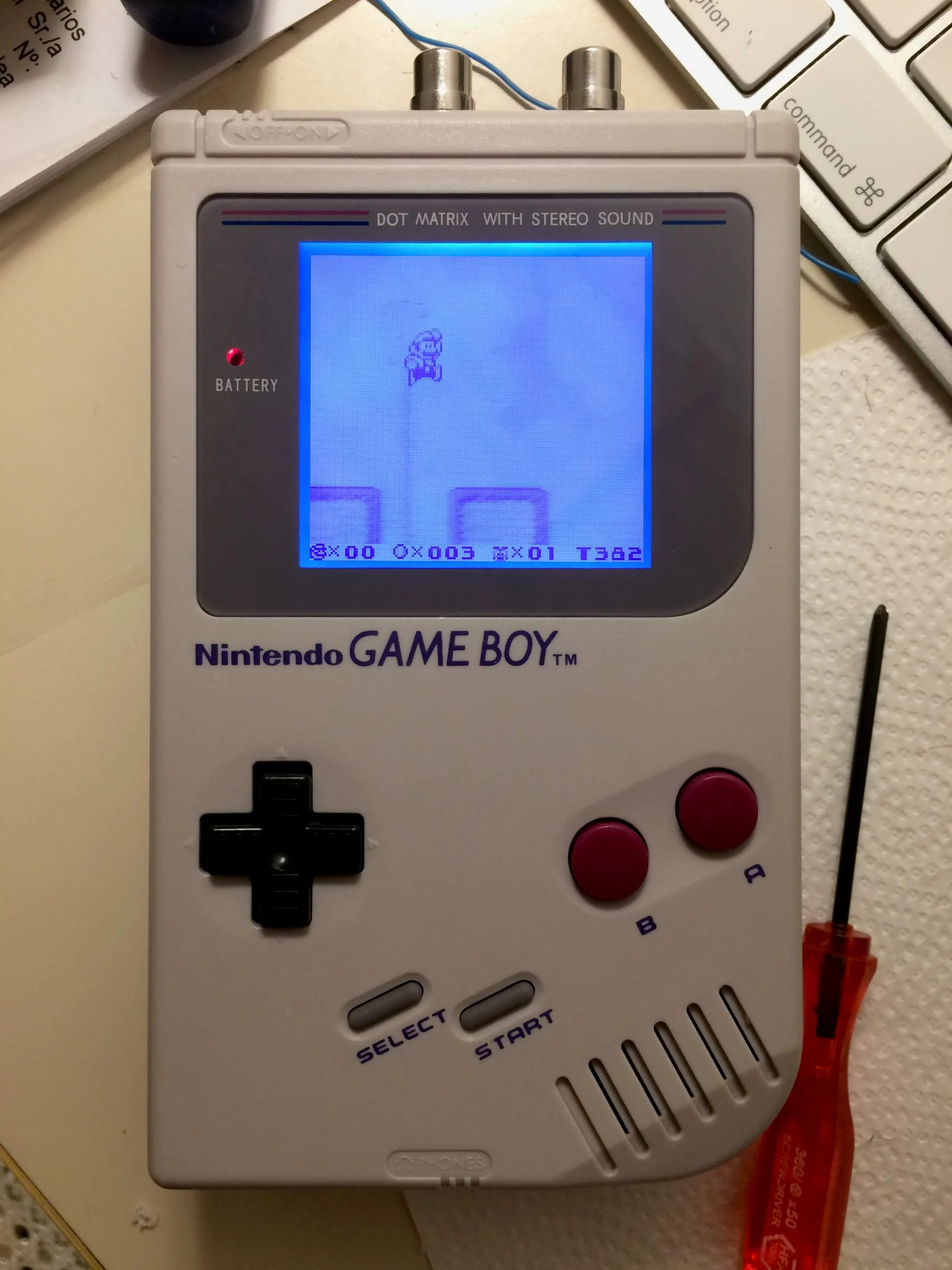 An assembled Game Boy in a clean pale grey shell, screen backlit pale blue showing Super Mario Land 2 in play. Two silver RCA jacks protrude from the top edge with a blue wire looped between them. A red screwdriver and a paper towel lie beside it on the desk.