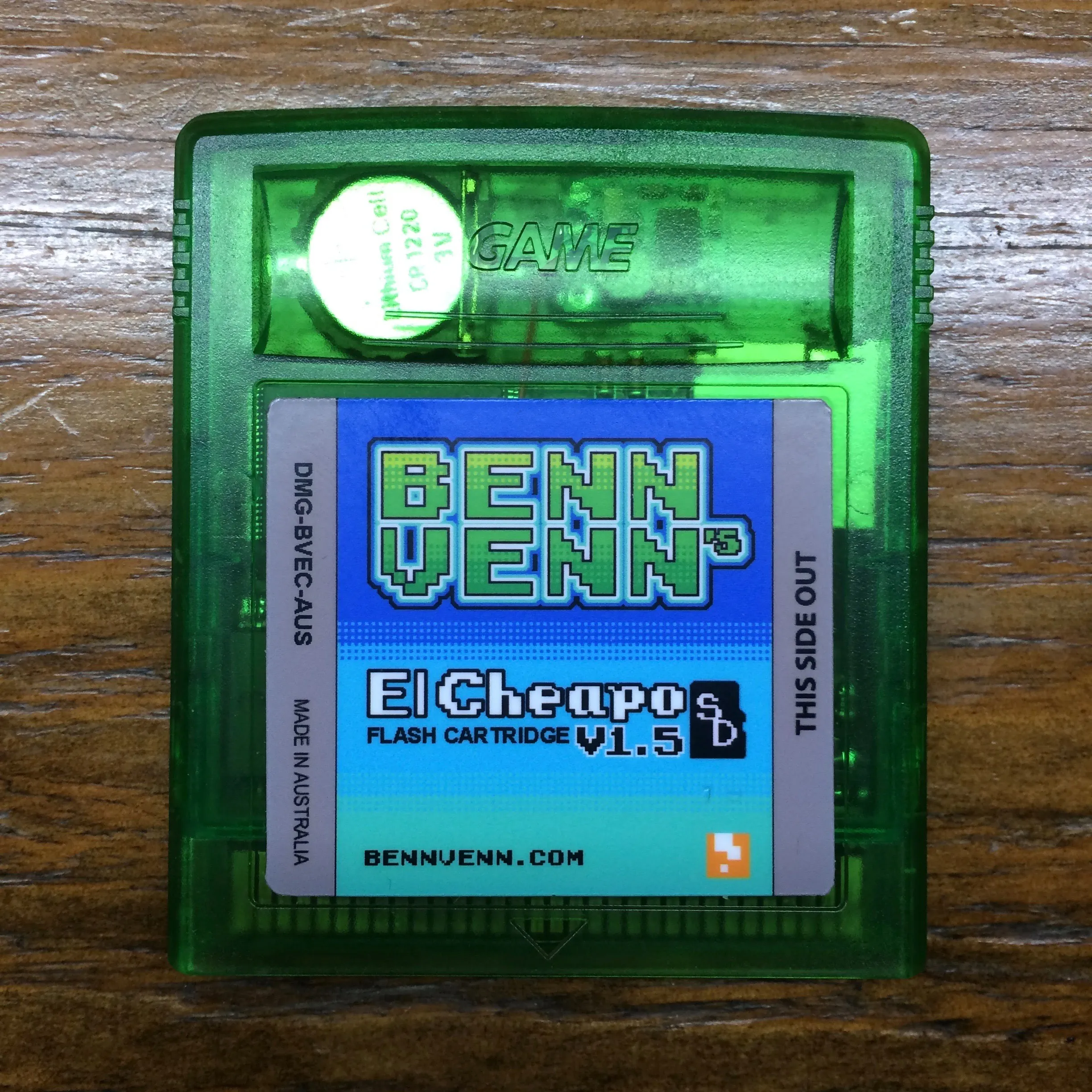 A translucent green Game Boy cartridge on a wooden surface, labelled BennVenn's El Cheapo SD Flash Cartridge v1.5, with a microSD slot and a coin cell visible through the shell. Small print reads DMG-BVEC-AUS and MADE IN AUSTRALIA.