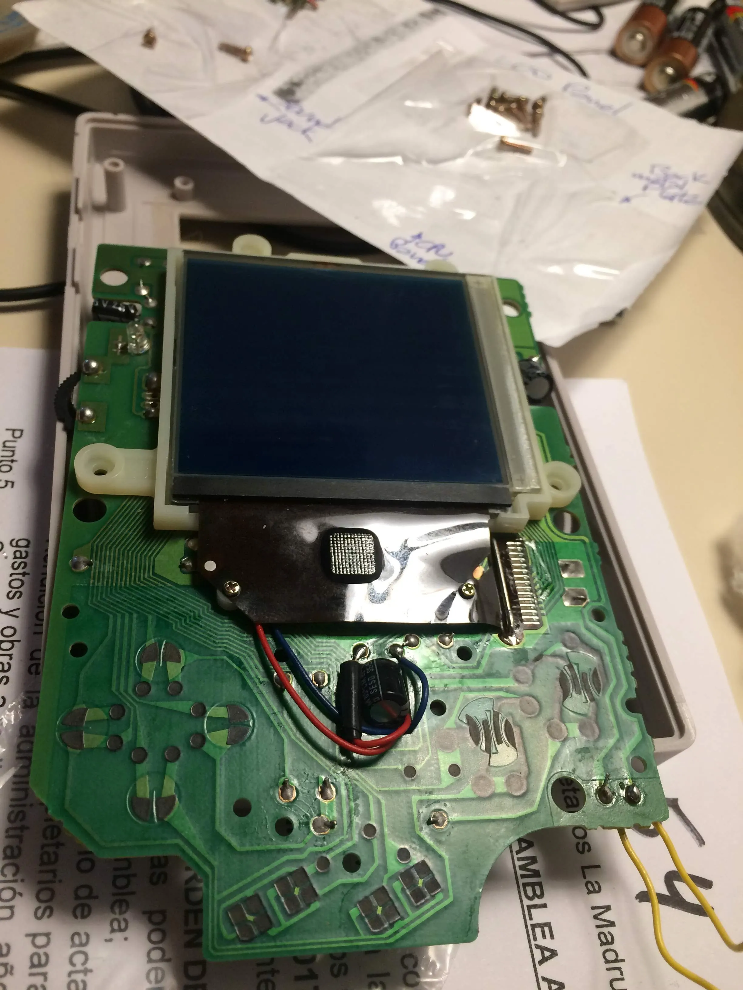 A Game Boy circuit board on a desk, LCD reattached and now deep navy blue with the polarising film removed. A red and a blue wire run from a small capacitor across the board towards the ribbon connector. A paper bag of labelled screws sits above.
