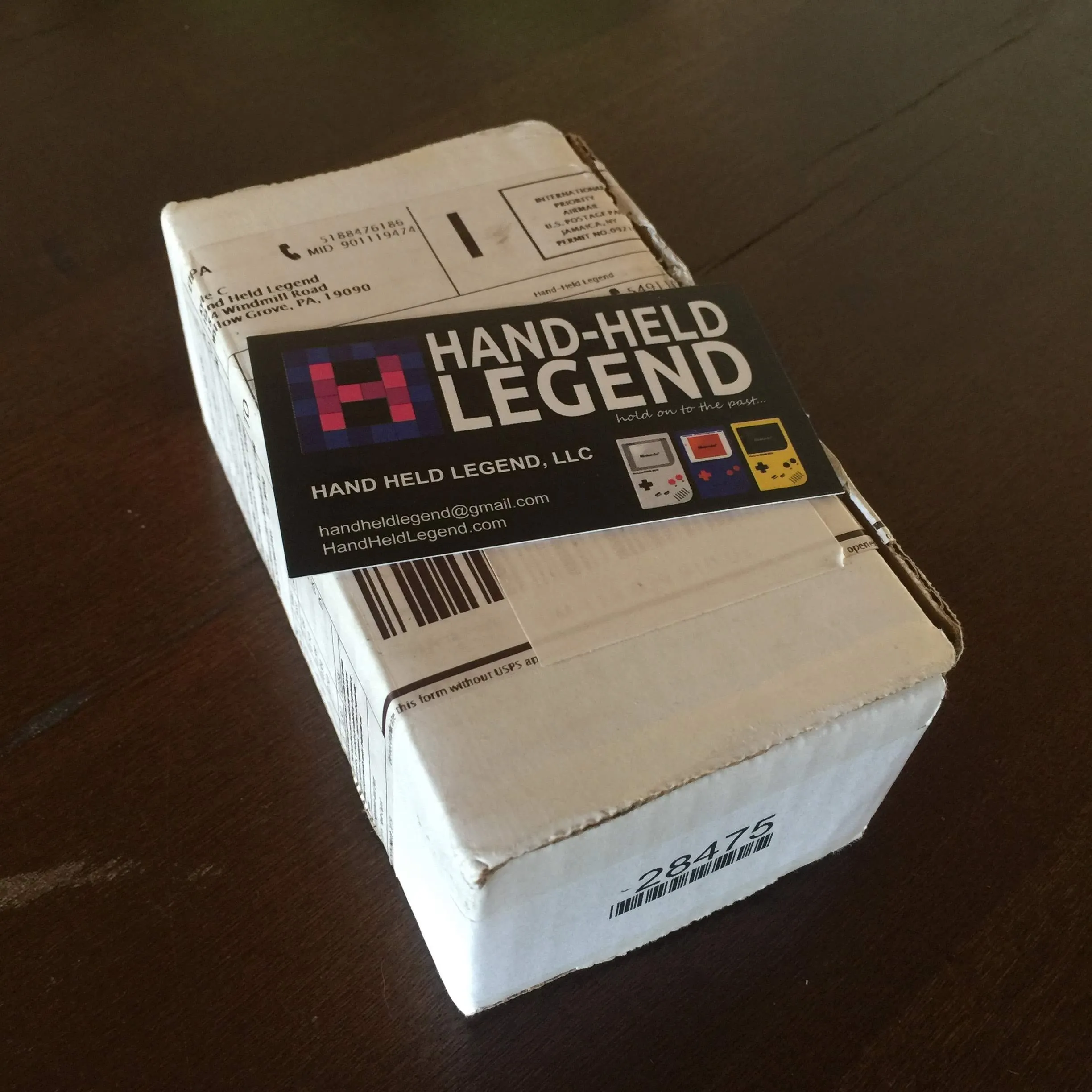 A small white cardboard box on a dark wooden table, sealed with a Hand-Held Legend sticker reading "hold on to the past" with three pixel-art Game Boys on it. International airmail markings and a customs form are visible along the top.
