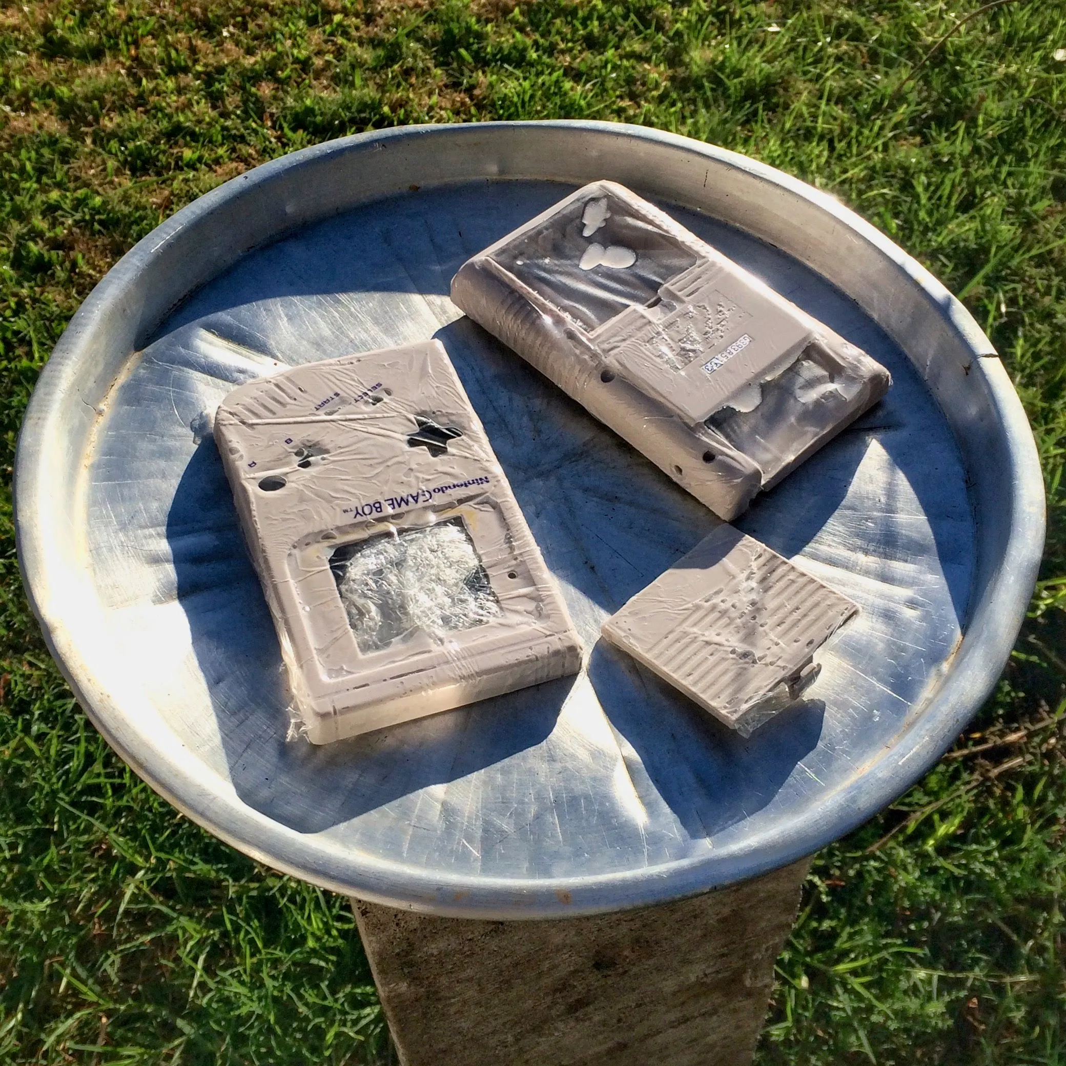 A round metal tray on a wooden post in a sunlit garden, holding three grey Game Boy shell pieces coated in white cream and wrapped tightly in clear stretch film. Bright green grass surrounds the post and the shadows are hard.