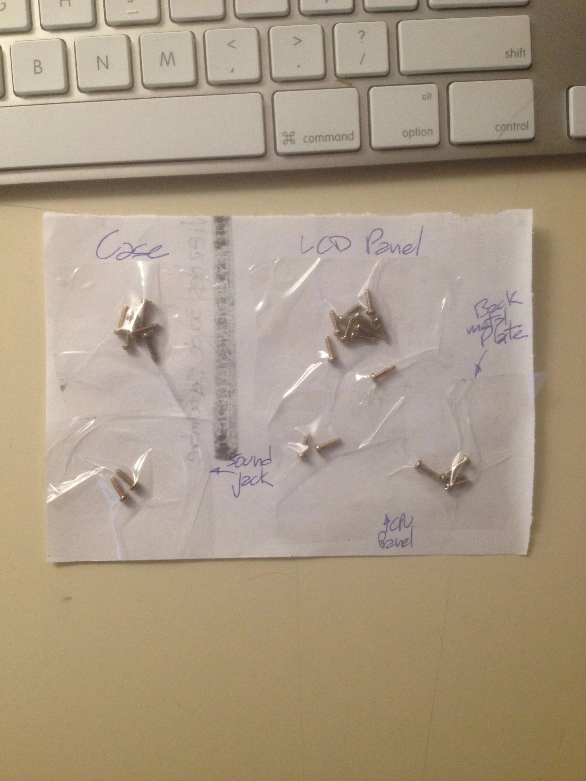 A sheet of white paper on a desk beside an Apple keyboard, divided into labelled zones in blue ballpoint: Case, LCD Panel, Sound Jack, Back Metal Plate. Small groups of silver screws are taped down flat inside each zone under strips of clear packing tape.