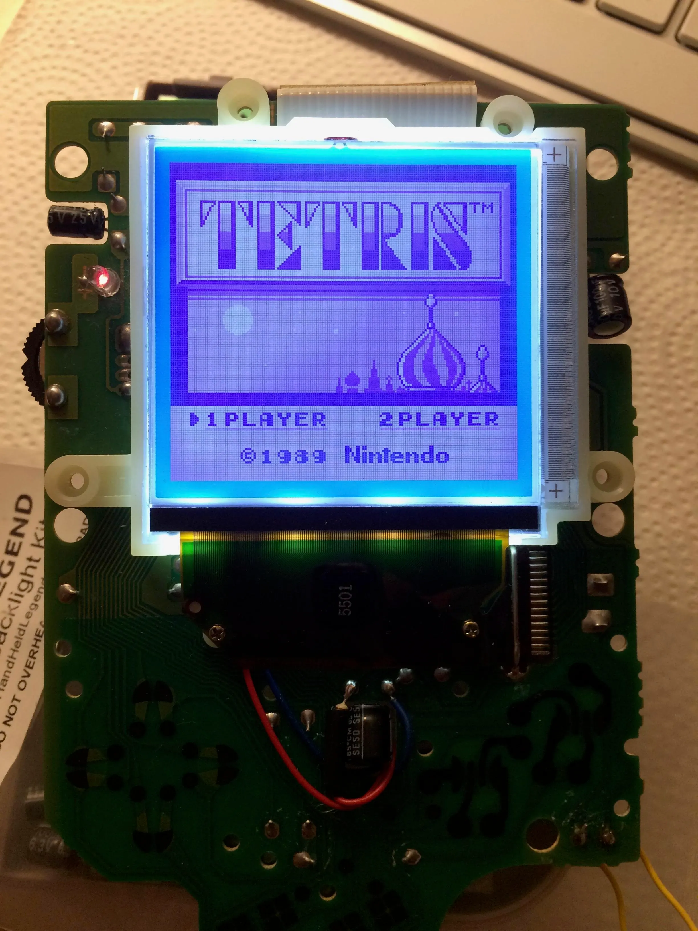 A Game Boy circuit board on a desk with the LCD lit bright blue, showing the Tetris title screen with the Kremlin illustration and 1 PLAYER and 2 PLAYER options. A small red LED glows at the left. A Hand Held Legend backlight kit instruction sheet is visible under the board.