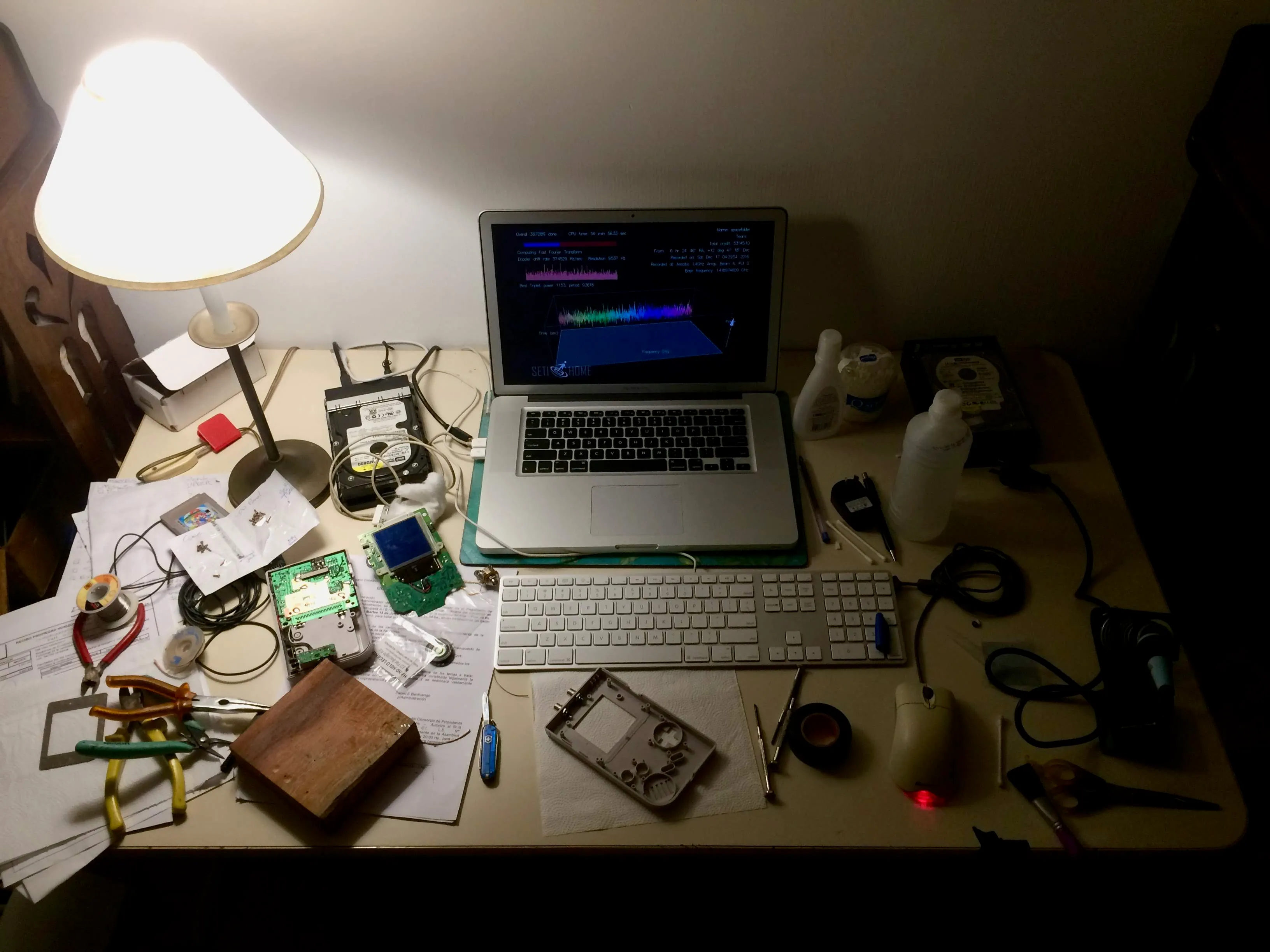 A desk at night lit by one lamp. A MacBook Pro sits in the middle running a SETI at home analysis screen with a waveform and spectrum plot. Around it: a Game Boy board with a blue LCD, the front shell on a paper towel, pliers, a solder spool, bottles of peroxide, a bare hard drive, a soldering iron and a screw map.