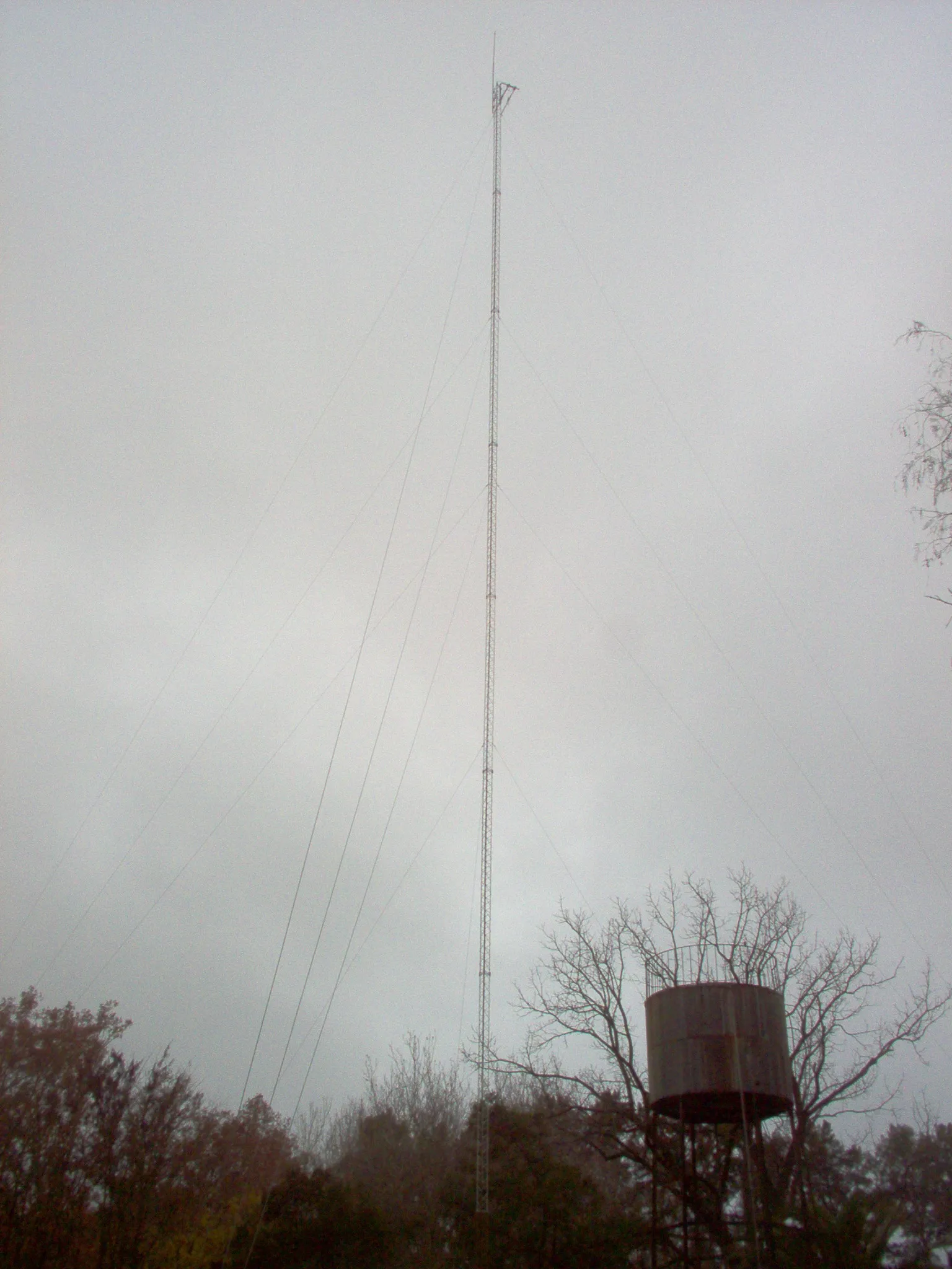 The tower at El Aduar photographed from the ground on an overcast day: a thin guyed lattice mast running the full height of the frame, six guy wires stretching down to their anchors, bare winter trees along the bottom and an elevated water tank at the right.