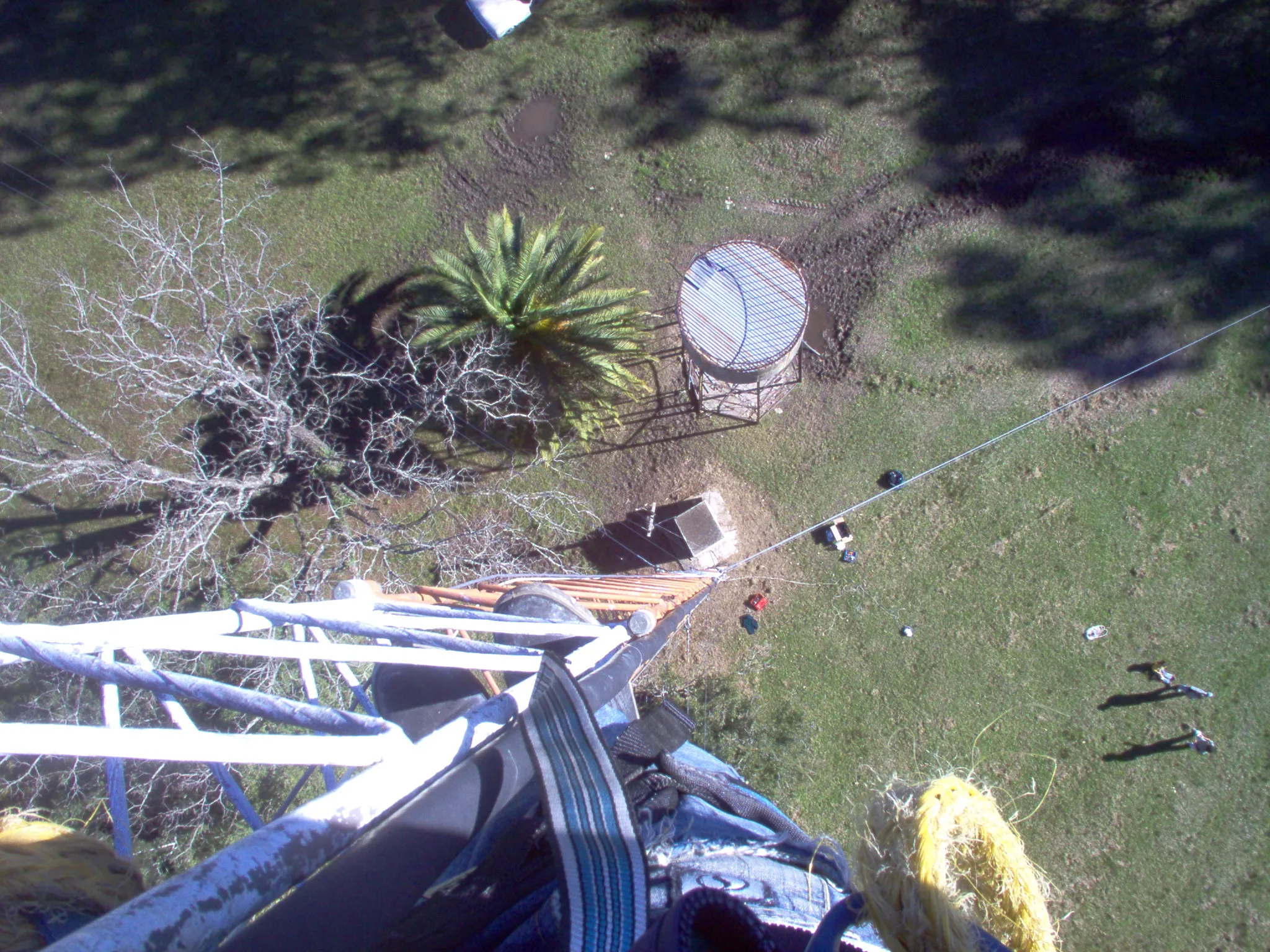 Looking straight down the tower from the working position: the photographer's own boots and the blue harness webbing fill the bottom of the frame, the white lattice and guy wires fall away below, and far down on the grass the elevated water tank reads as a small circle while two people stand as specks casting long shadows.