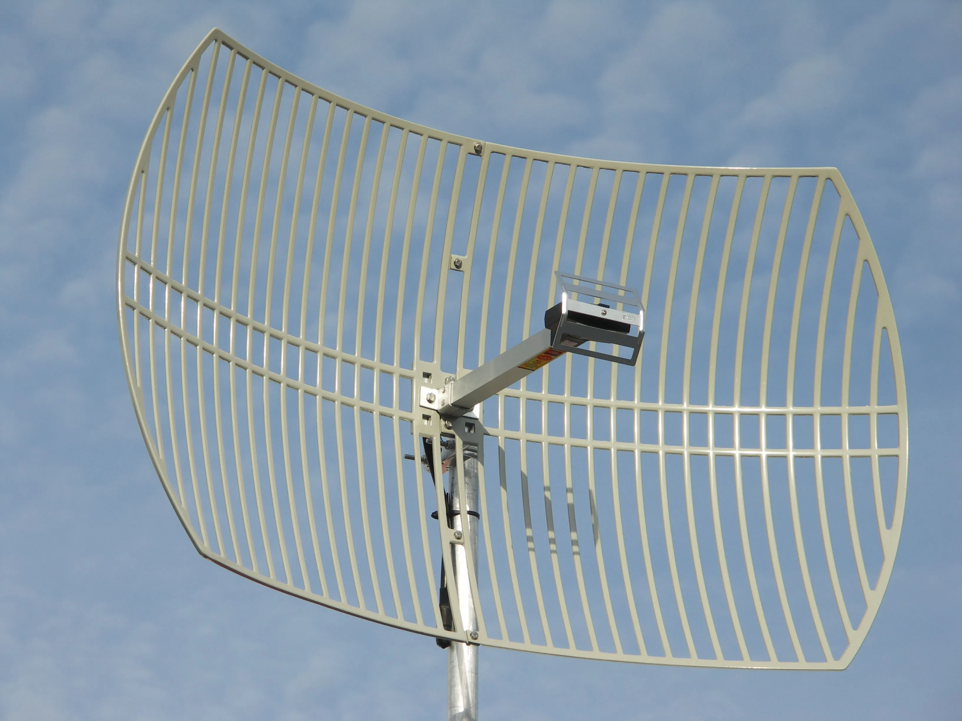 A grid parabolic antenna against a blue sky: a curved rectangular reflector made of pale aluminum bars on a horizontal frame, with a small boxed feed on an arm at its focus, clamped to a galvanized mast.